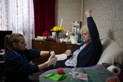 A women sitted in a sofa is doing exercises, hand raised and follow the advices of HI physio Maria who is sitted next to her doing the same with her hands