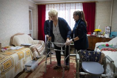 Antonina is walking thanks to a mobility aid in the small room of the appartement where we can see a toilet chair a sofa and a table, helped by Maria the physio on her right